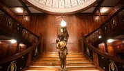 An ornate bronze statue of a figure holding a lamp stands at the base of a grand wooden staircase with detailed craftsmanship.
