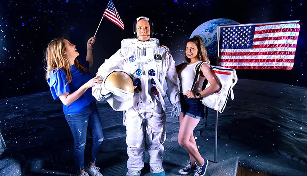 Two people are posing with a life-size astronaut mannequin on a staged moon surface complete with a backdrop of space and an American flag