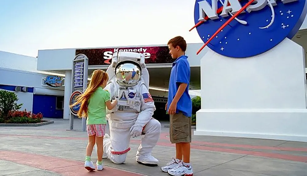 A person in an astronaut suit is kneeling to interact with a young girl while a boy stands nearby in front of the Kennedy Space Center sign