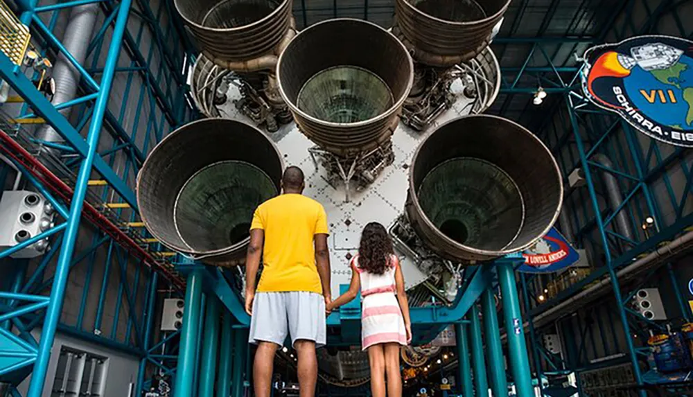 A man and a young girl are standing in front of the massive engine nozzles of a Saturn V rocket on display possibly at a space museum