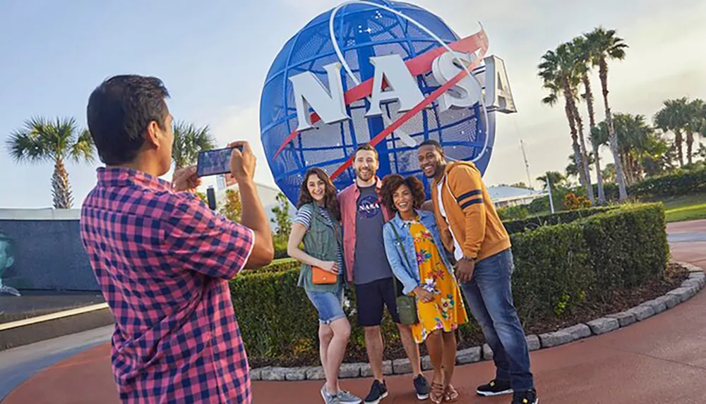 A group of four smiling people pose for a photo in front of a large NASA emblem while another person takes their picture with a smartphone