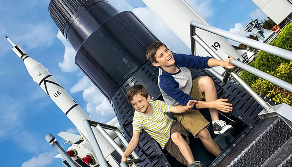 Two children are smiling and posing for a photograph with space rockets in the background likely at a space-themed park or museum