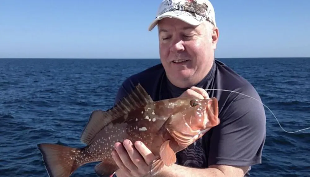 A smiling man is holding a large fish on a sunny day with the ocean in the background