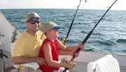 A smiling man in sunglasses and yellow attire guides a young boy in a red sleeveless shirt and cap as they fish together on a boat at sea.