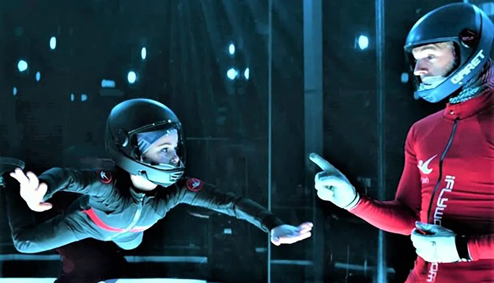 Two individuals equipped with helmets and suits appear to be in an indoor skydiving facility with one person gesturing towards the other as if giving instructions or guidance