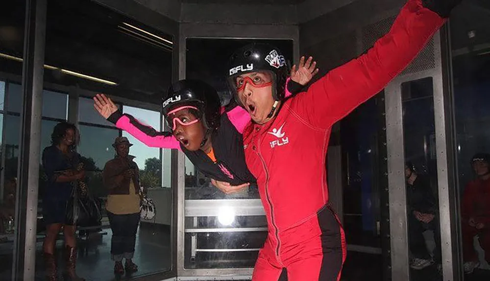 Two people are indoor skydiving in a wind tunnel with one person in a pink and black suit flying with the assistance of an instructor in a red suit while spectators watch from outside