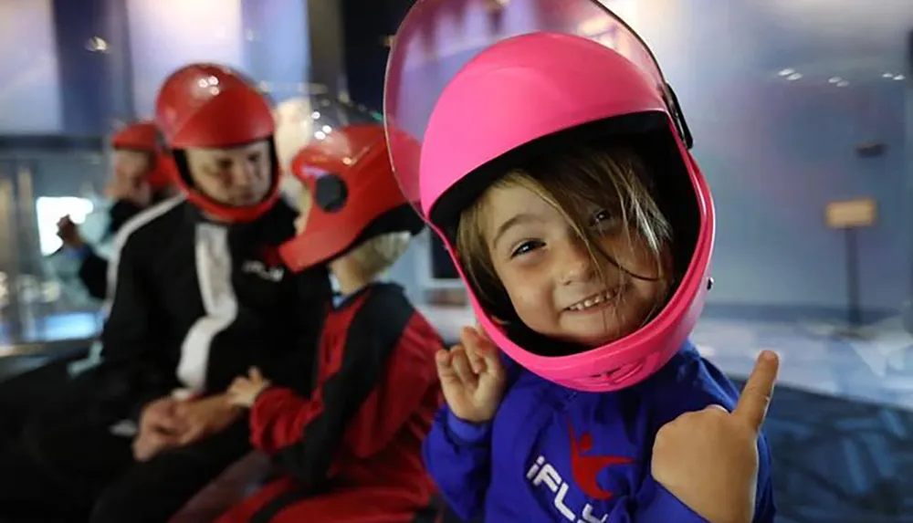 A cheerful child wearing a large pink helmet gives a thumbs up likely excited about an indoor skydiving experience as suggested by the outfit and surrounding participants with helmets