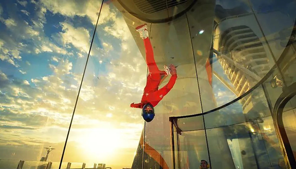 A person in a red suit is performing an indoor skydiving maneuver inside a vertical wind tunnel set against a backdrop of a sunset sky