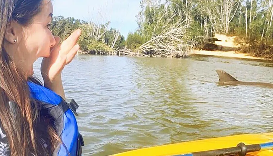 A person is seemingly blowing a kiss to a shark fin above the water while kayaking.