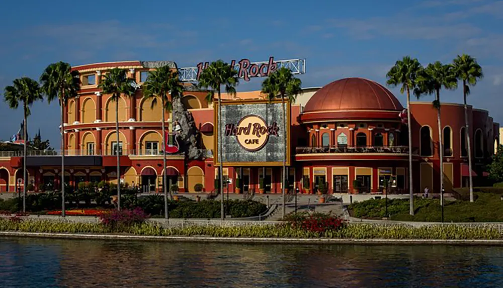 This image shows the exterior of a Hard Rock Cafe building with its iconic logo and distinctive architecture situated near a body of water under a clear sky with palm trees around