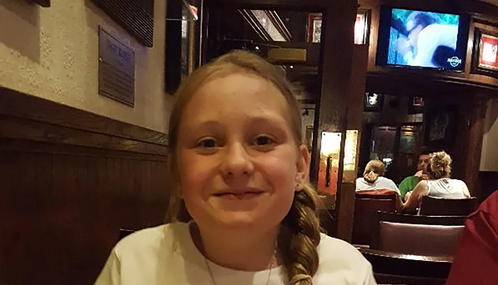 A smiling young girl with a side braid is sitting in what appears to be a pub or a restaurant with a television screen and other patrons in the background