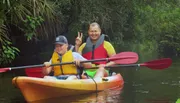 Two people are smiling and posing with peace signs while kayaking together in a verdant river setting.