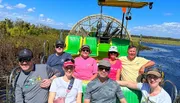 A group of smiling people is posing for a photo on an airboat during a sunny day, likely on a marsh or swamp tour.