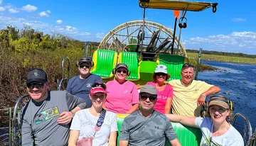 A group of smiling people is posing for a photo on an airboat during a sunny day, likely on a marsh or swamp tour.