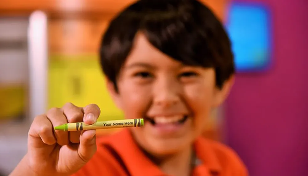 A smiling child is holding up a green crayon with the placeholder text Your Name Here on its label with a colorful blurred background