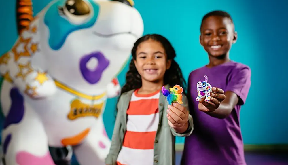 Two children are smiling as they proudly display colorful toys in their hands with a whimsically decorated statue of a dog in soft focus behind them