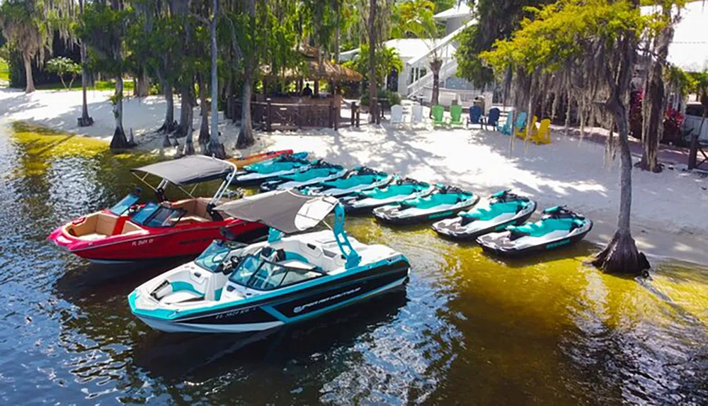 The image shows a variety of watercraft including boats and jet skis moored near a sandy beach area with trees and some colorful beach chairs suggesting a recreational water activity spot