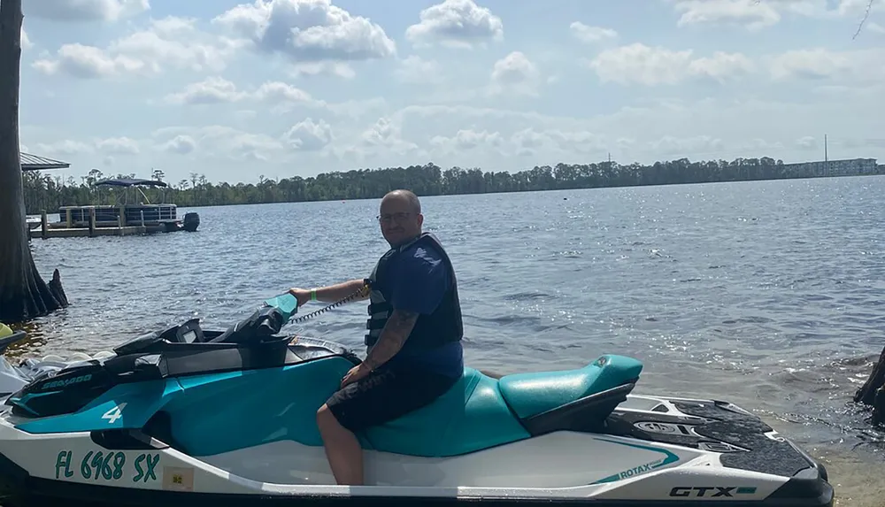 A person is seated on a teal and white jet ski near the shore of a lake with a pontoon boat and trees in the background under a partly cloudy sky