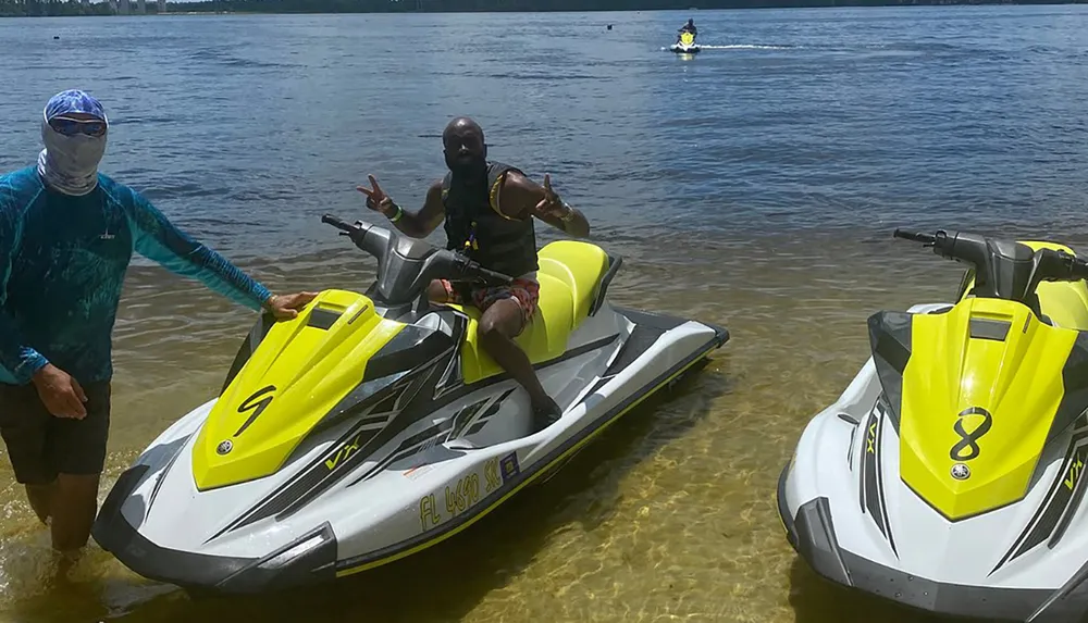 Two people are enjoying a sunny day with jet skis by the lakeshore with one person on a jet ski making a peace sign and another standing beside a jet ski