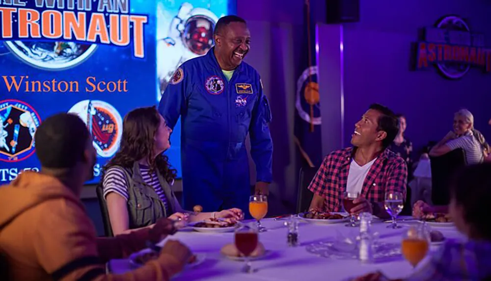 An astronaut is sharing a jovial moment with guests at a Dine with an Astronaut event as they enjoy their meal and seem delighted by the interaction