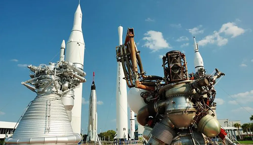 The image features a close-up of rocket engines in the foreground with a collection of historic rockets on display under a blue sky likely at a space museum or exhibit
