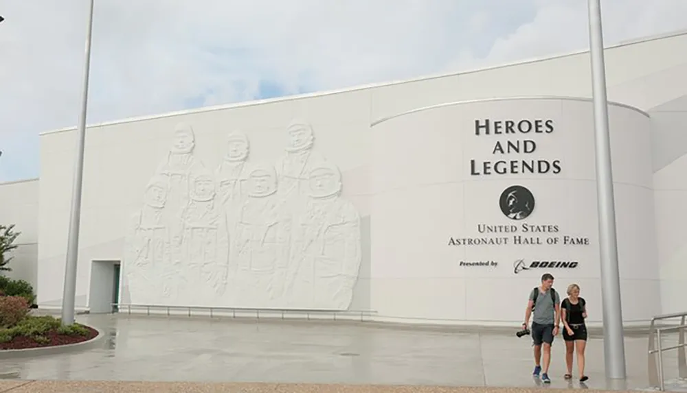 The image shows two people walking past the Heroes and Legends building which houses the United States Astronaut Hall of Fame featuring a large relief of astronauts on its facade