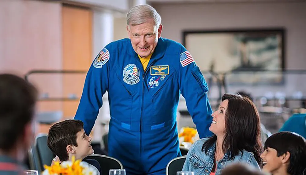 An individual in a NASA astronaut suit is leaning over a table to interact with smiling people seated around it