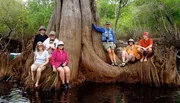 A group of people in outdoor attire are posing cheerfully around a large tree with protruding roots by a body of water.