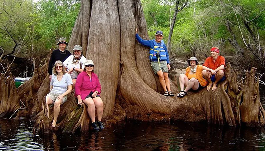 A group of people in outdoor attire are posing cheerfully around a large tree with protruding roots by a body of water.