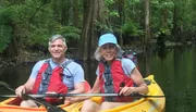 Two people are smiling while kayaking together on a calm river surrounded by greenery.