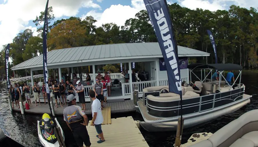 A group of people gathered on a dock by a waterway, with pontoon boats nearby and promotional flags fluttering in the breeze.