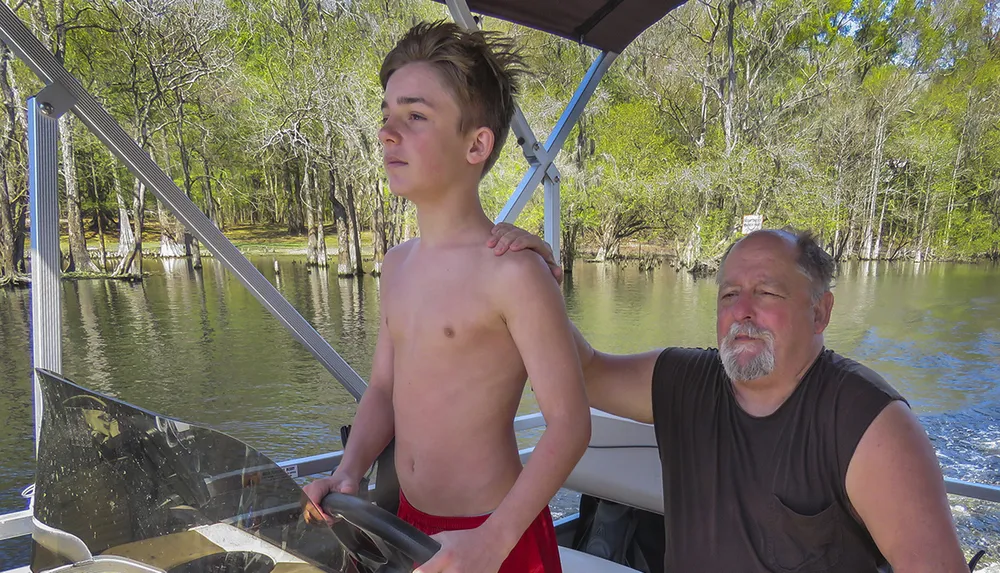 A young person is at the helm of a pontoon boat while an older individual sits beside them both enjoying a sunny day on the water amidst a wooded area
