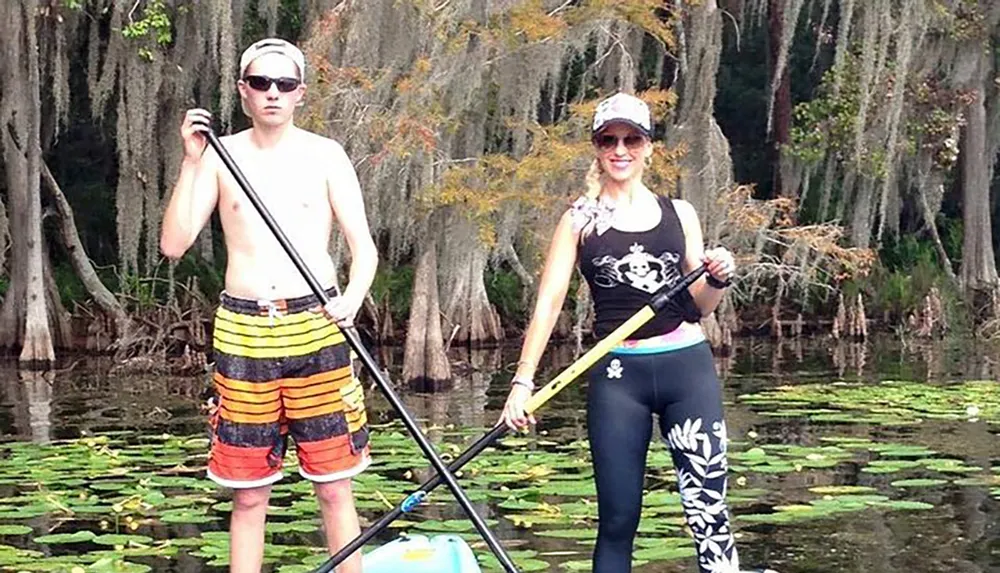 A man and a woman are standing on paddleboards in a calm waterway with lily pads surrounded by trees with hanging Spanish moss