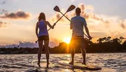 Two people are stand-up paddleboarding on calm water against a beautiful backdrop of a setting sun.