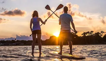 Two people are stand-up paddleboarding on calm water against a beautiful backdrop of a setting sun.