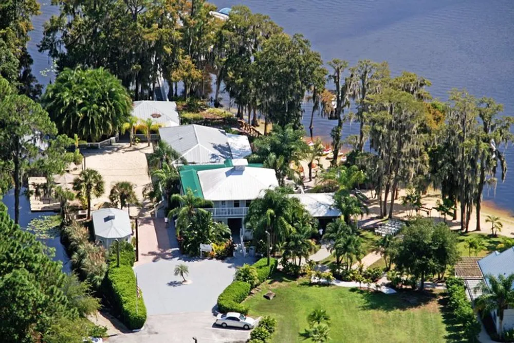 The image is an aerial view of a large luxurious estate with a green roof surrounded by lush greenery situated next to a body of water