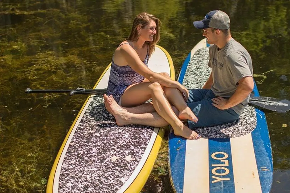 A man and a woman are sitting on paddleboards on a calm body of water facing each other in conversation
