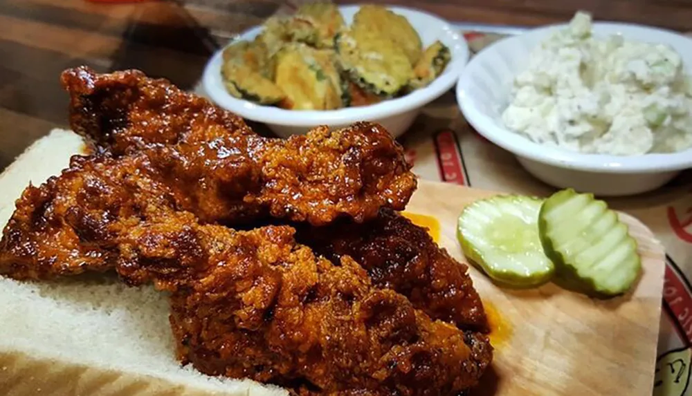 The image shows a serving of crispy fried chicken on white bread with pickles accompanied by sides of coleslaw and fried pickles in the background
