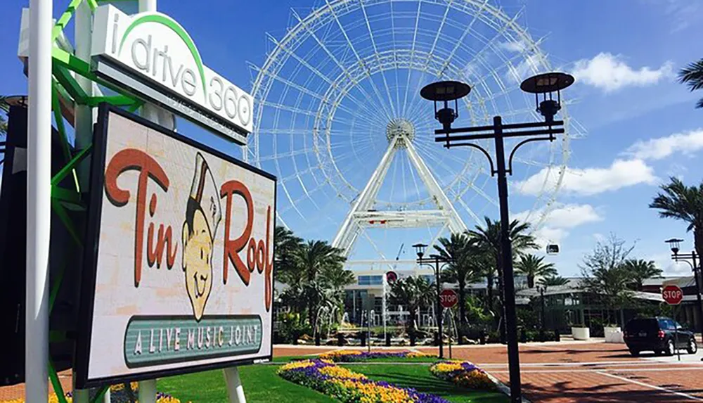 The image shows a sunny day at a leisure complex with the sign Tin Roof - A Live Music Joint in the foreground and a large observation wheel in the background