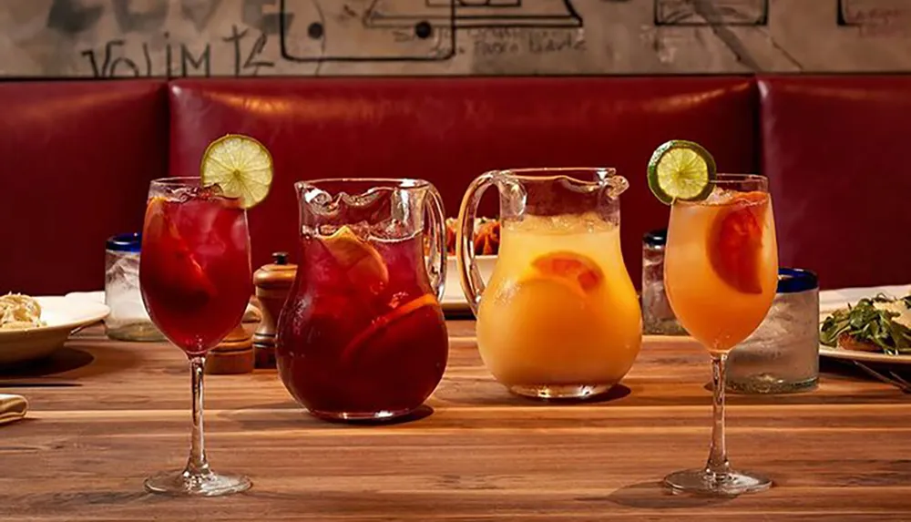 The image shows a refreshing scene with two pitchers of colorful beverages one red and one yellow accompanied by filled glasses garnished with lime slices all set on a wooden table against a red leather booth backdrop