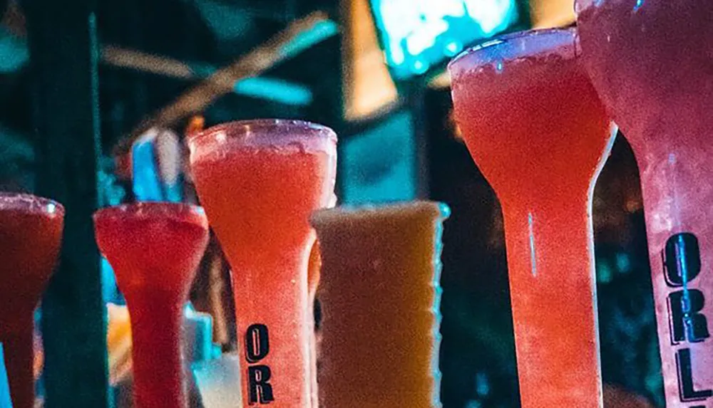 A row of colorful frozen cocktails is displayed at a bar with a blurred background emphasizing the vibrant nightlife atmosphere