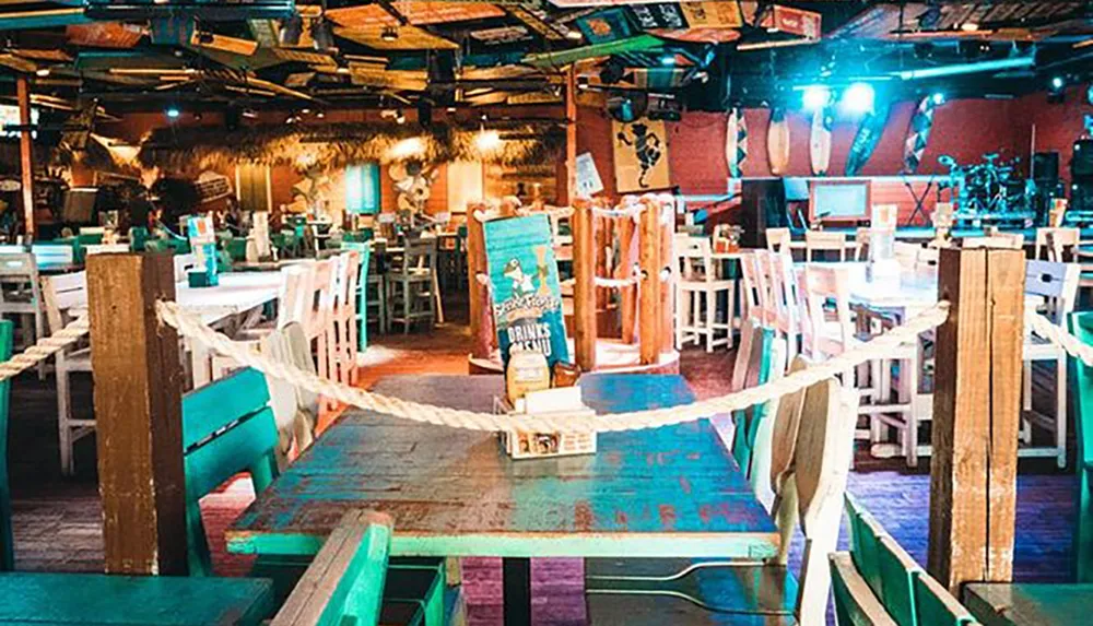 The image shows a colorful beach-themed bar with empty chairs vibrant decorations and a stage set up for live music