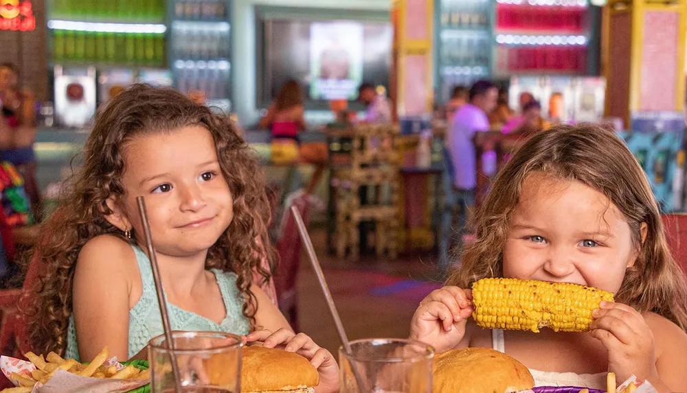 Two young girls enjoy a meal at a colorful restaurant with one happily biting into a cob of corn