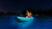 Two people are kayaking at night on a bioluminescent bay under a starry sky.