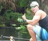 A man is giving a thumbs-up while sitting on a boat seemingly unaware of a large black animal possibly a bear in the background among the foliage