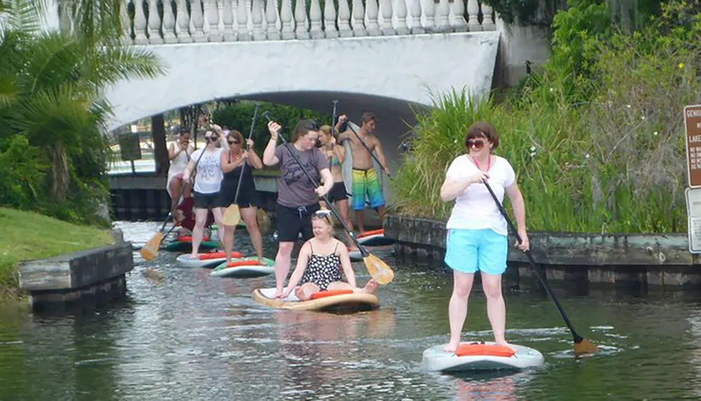 A group of people are stand-up paddleboarding on a waterway with a bridge in the background