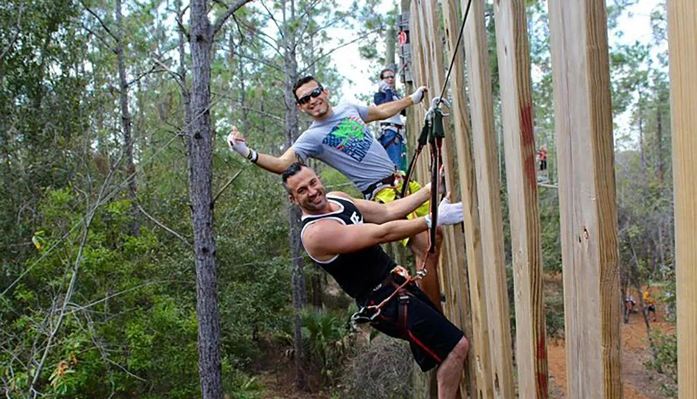 Three people are climbing a tall wooden structure outdoors secured with harnesses and ropes displaying expressions of enjoyment and adventure