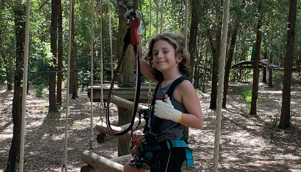 A smiling child gives a thumbs up while wearing safety gear for an outdoor adventure or ropes course in a wooded area
