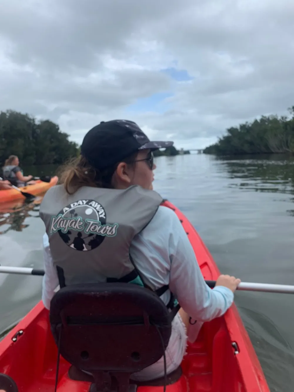 A person is seen from behind while kayaking on a cloudy day wearing a cap and a life vest labeled A Day Away Kayak Tours