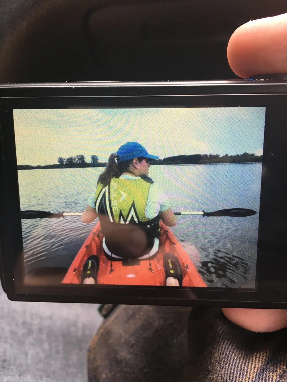 A person is kayaking on a calm body of water viewed over the shoulder from a cameras display screen